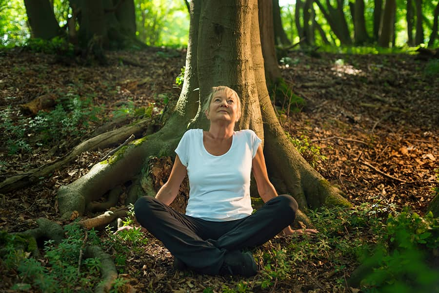 Image of a woman sitting cross-legged. She is in a forest and sitting underneath a tree. She looks calm.